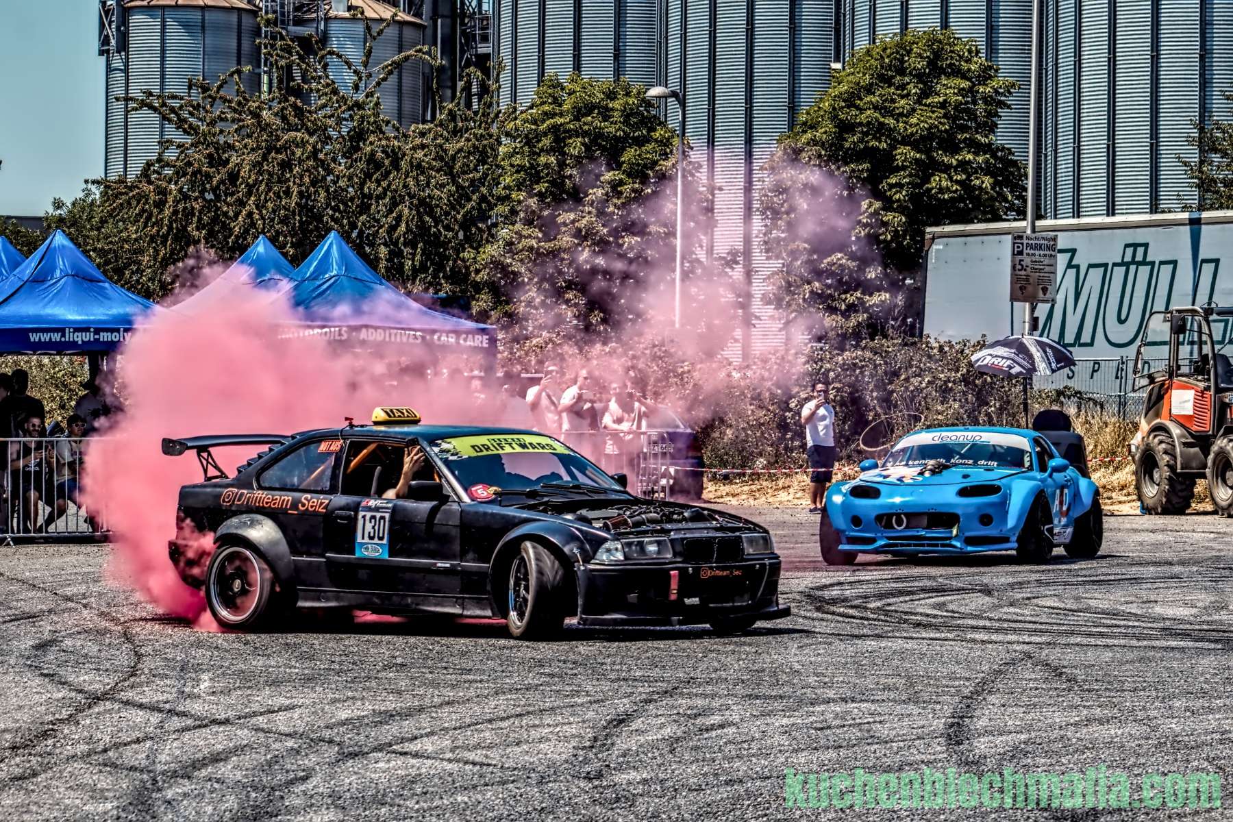Schwarzes BMW Drift-Auto erzeugt pinken Reifenrauch auf einem asphaltierten Platz vor großen Silos.