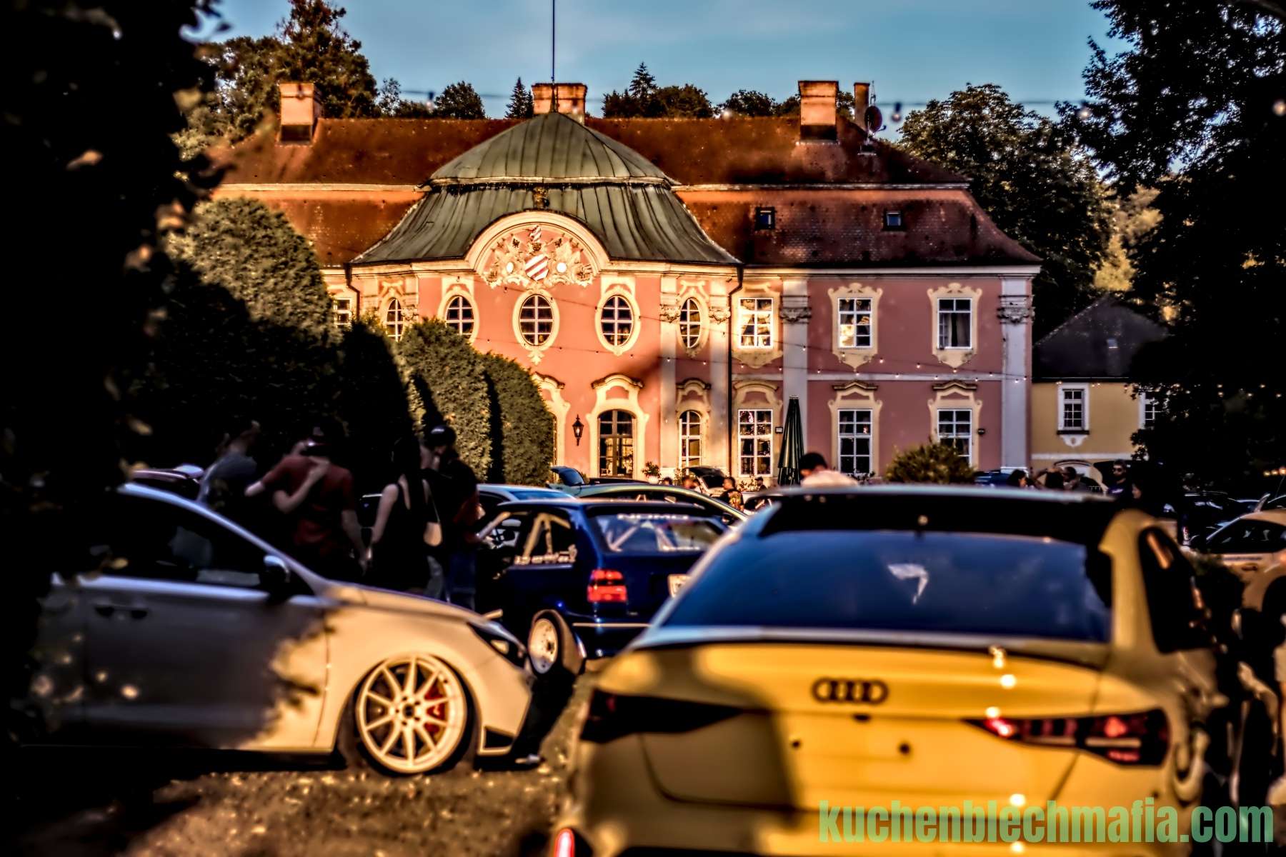 Tiefergelegte Autos parken vor einem barocken Schloss bei Abenddämmerung.