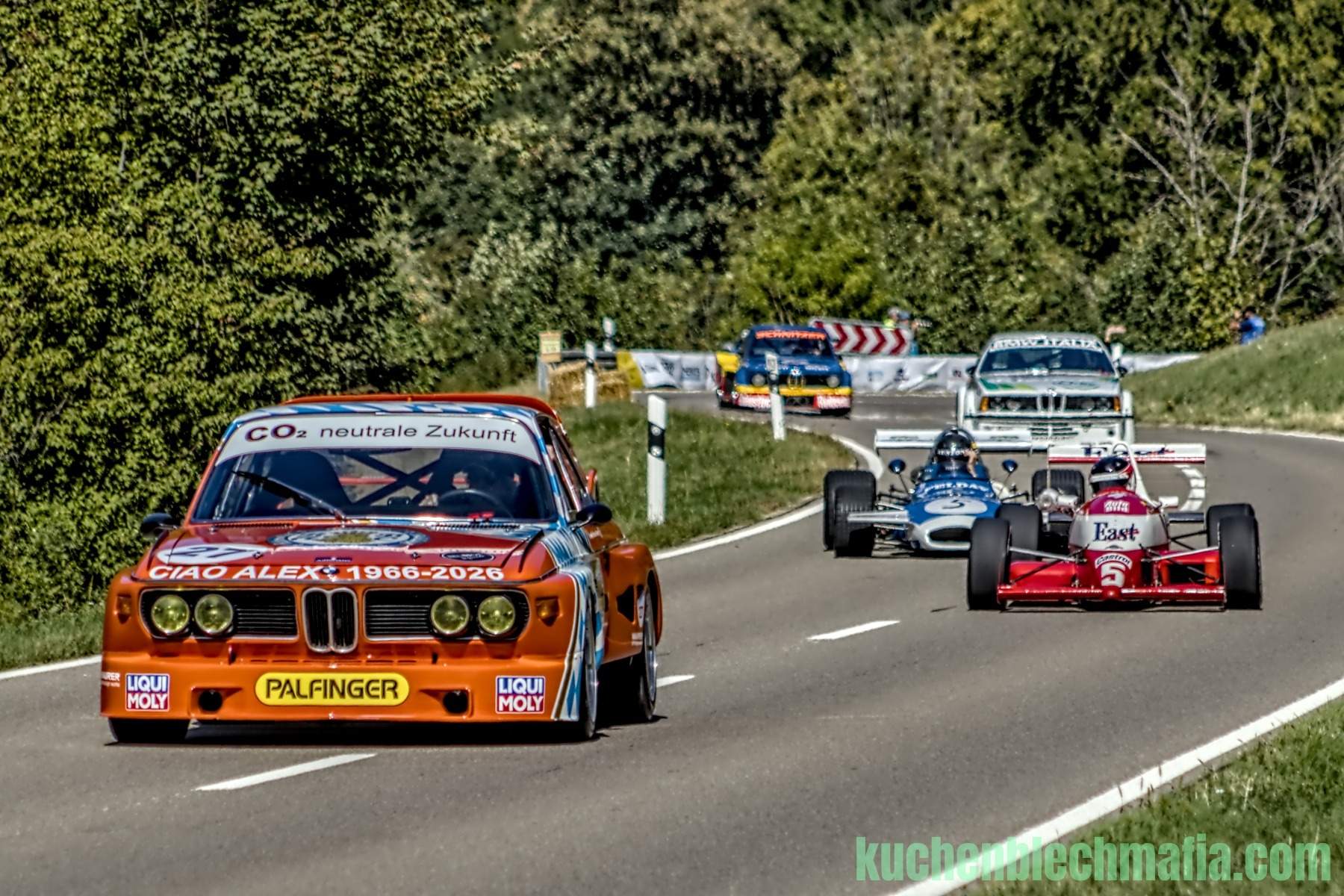 Orange BMW 3.0 CSL Rennwagen fährt vor rotem Formel-Rennwagen auf einer Landstraße.