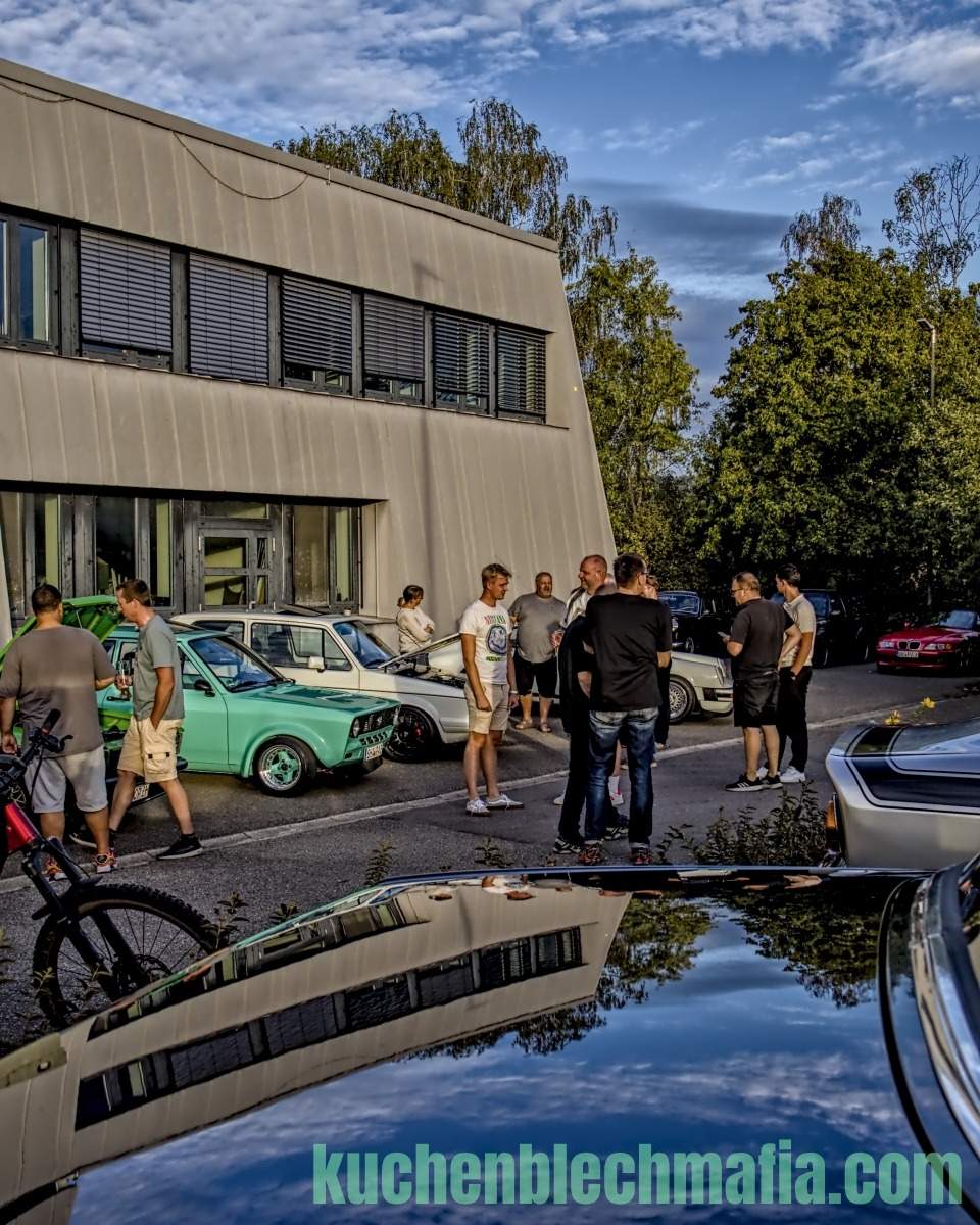 Männer stehen auf einem Parkplatz vor einem Gebäude und unterhalten sich neben einem mintgrünen Trabant.