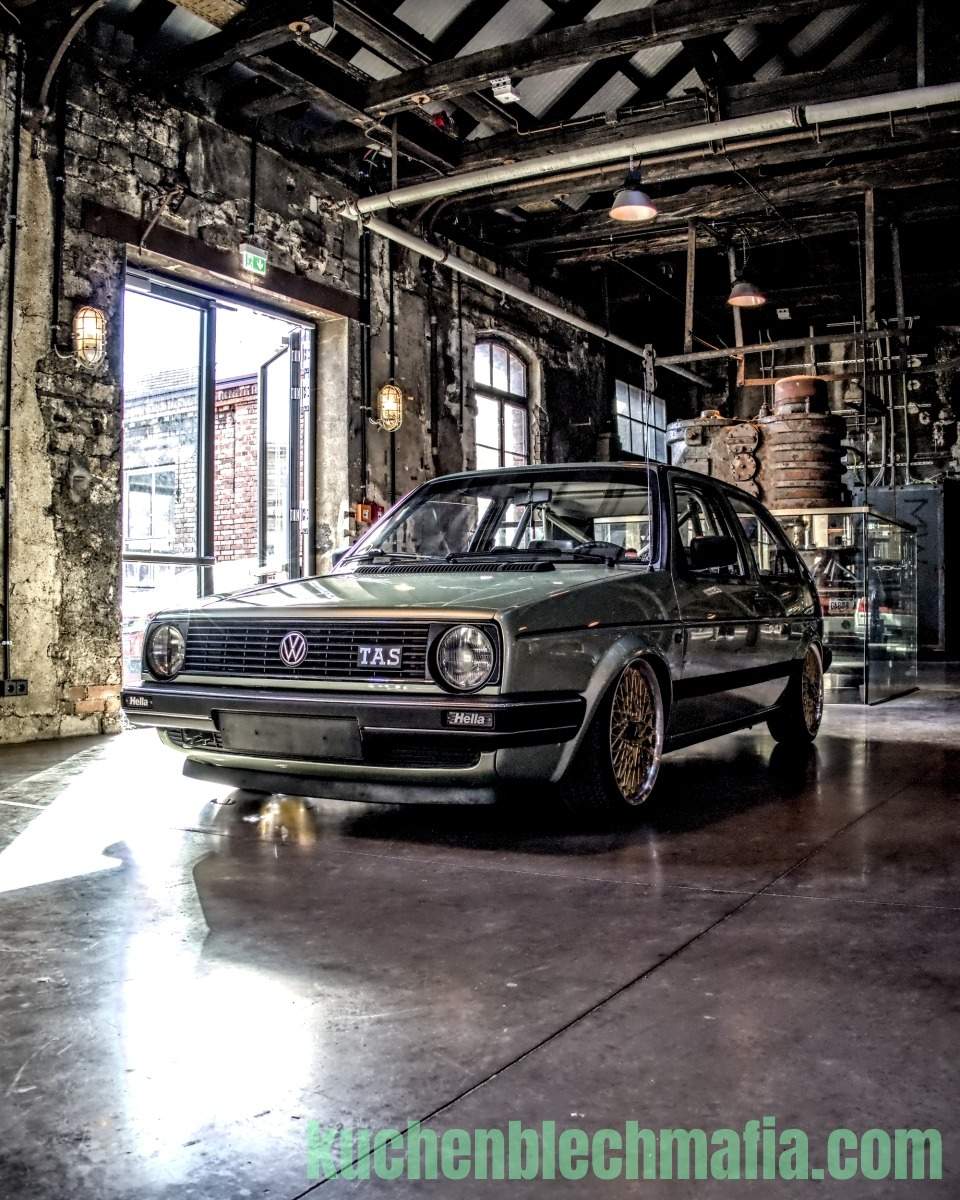 Ein modifizierter olivgrüner VW Golf II mit goldenen Speichenfelgen steht in einer industriellen Lagerhalle.