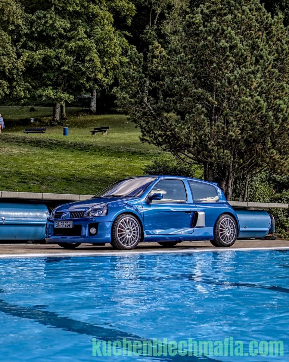 Leuchtend blauer Renault Clio V6 Sportwagen parkt an einem Pool vor grüner Parklandschaft.
