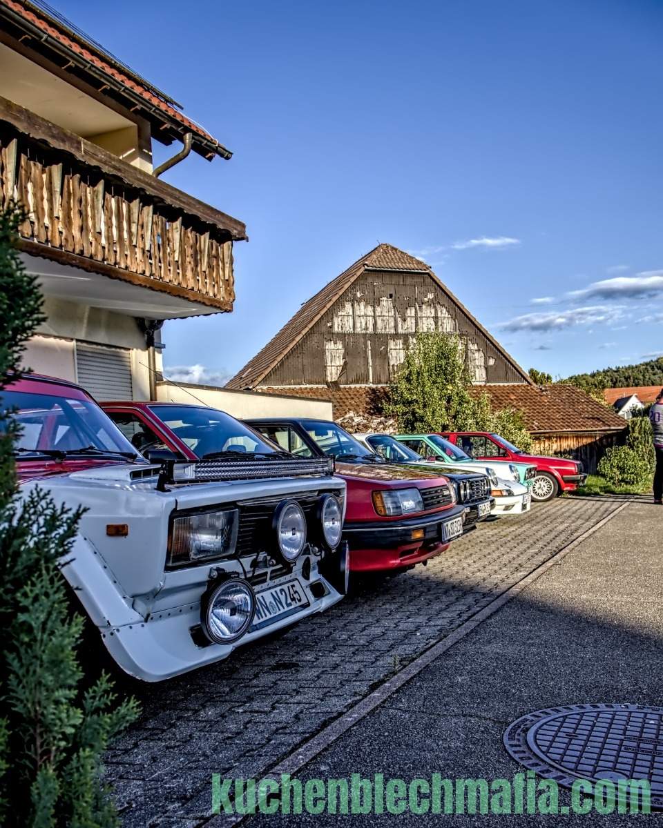 Weißer Oldtimer mit Zusatzscheinwerfern parkt vor weiteren Autos und einem Holzhaus