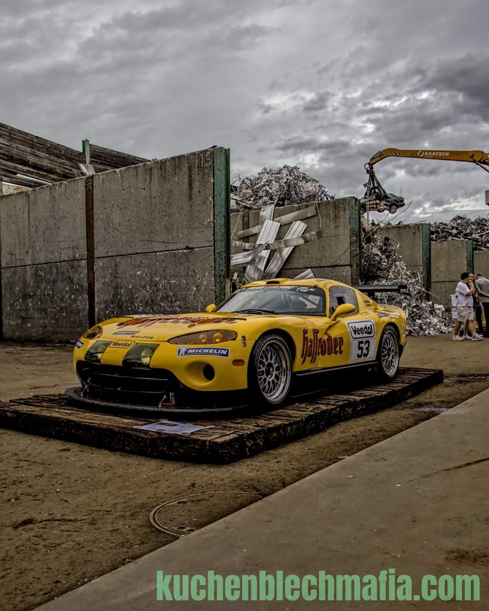 Gelber Dodge Viper mit Werbeaufschrift auf Holzplattform vor Betonwänden und Schrottplatz.