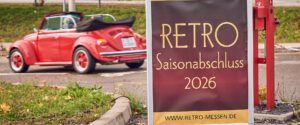 Roter VW Käfer Cabrio mit schwarzem Verdeck auf einer Straße, daneben ein Banner „RETRO Saisonabschluss 2026“ und ein roter Hydrant.