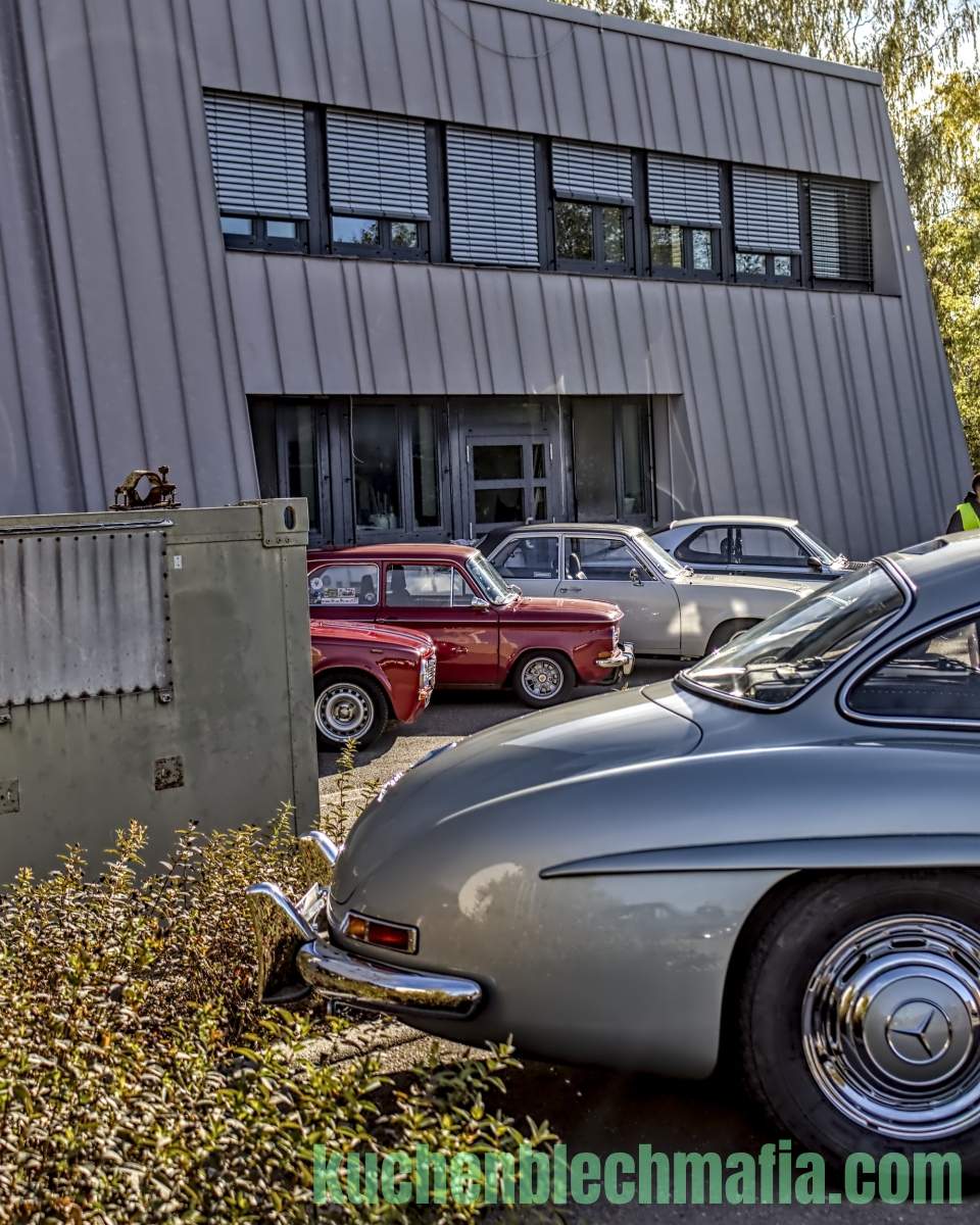 Silberner Mercedes 300 SL Flügeltürer parkt vor grauem Industriegebäude neben roten und weißen Oldtimern.