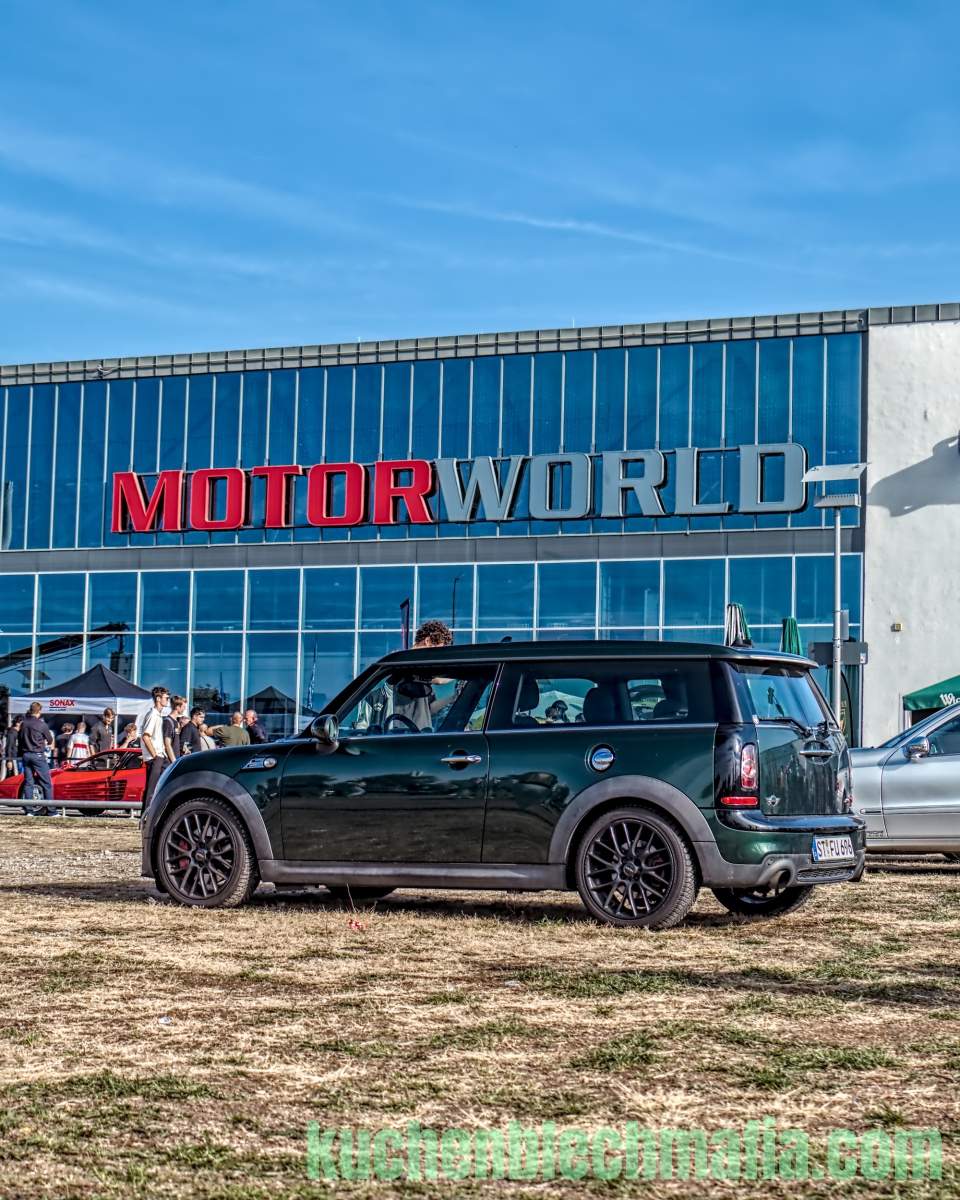 Dunkelgrüner Mini Clubman steht auf einer trockenen Wiese vor dem Gebäude von Motorworld.